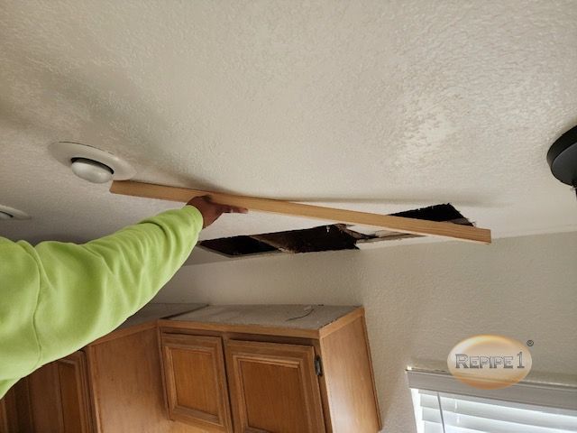 Person in a green shirt holding a wooden plank near a ceiling with exposed cutouts, showing ongoing repair or maintenance work above kitchen cabinets.