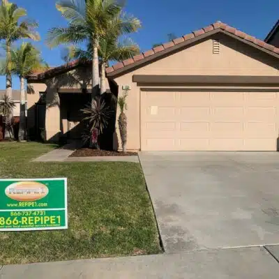 House with a sign for a plumbing service on the lawn, featuring a beige garage door and palm trees in the yard.