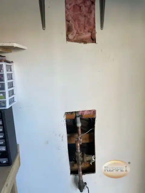 Wall with cutouts revealing plumbing pipes and insulation, indicating ongoing repair or maintenance work in a residential space.