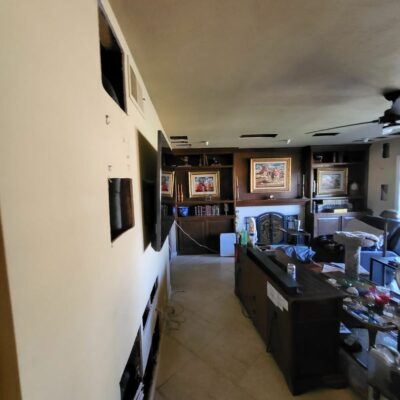 Living room with multiple cutouts in the wall and ceiling exposing internal structures, indicating active renovation or repair work, with furniture and decorations in the background.