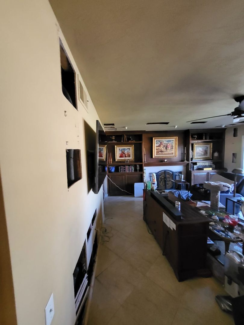 Living room with multiple cutouts in the wall and ceiling exposing internal structures, indicating active renovation or repair work, with furniture and decorations in the background.
