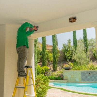 Worker on a ladder fixing a ceiling on a patio near a pool.