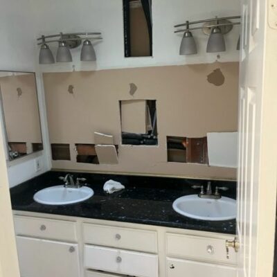 Partially demolished bathroom with dual sinks and broken drywall exposing internal structures, showing renovation progress.