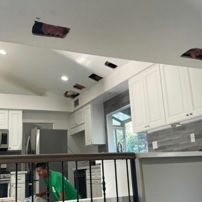 Modern kitchen with ceiling cutouts revealing insulation, indicating ongoing maintenance or renovation, while a worker in a green shirt attends to tasks below.