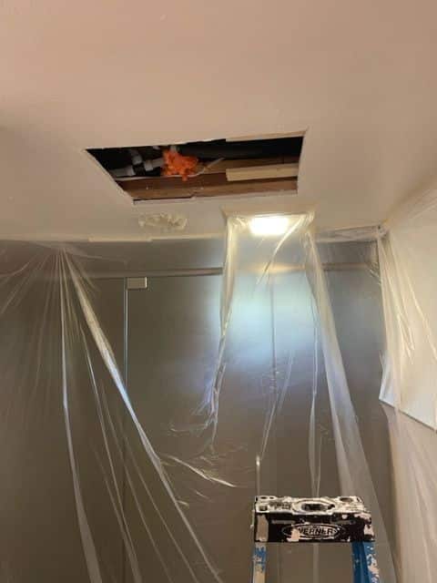 Ceiling with a large cutout exposing pipes and internal structures, covered with plastic sheeting to protect the area, indicating active renovation or repair work.